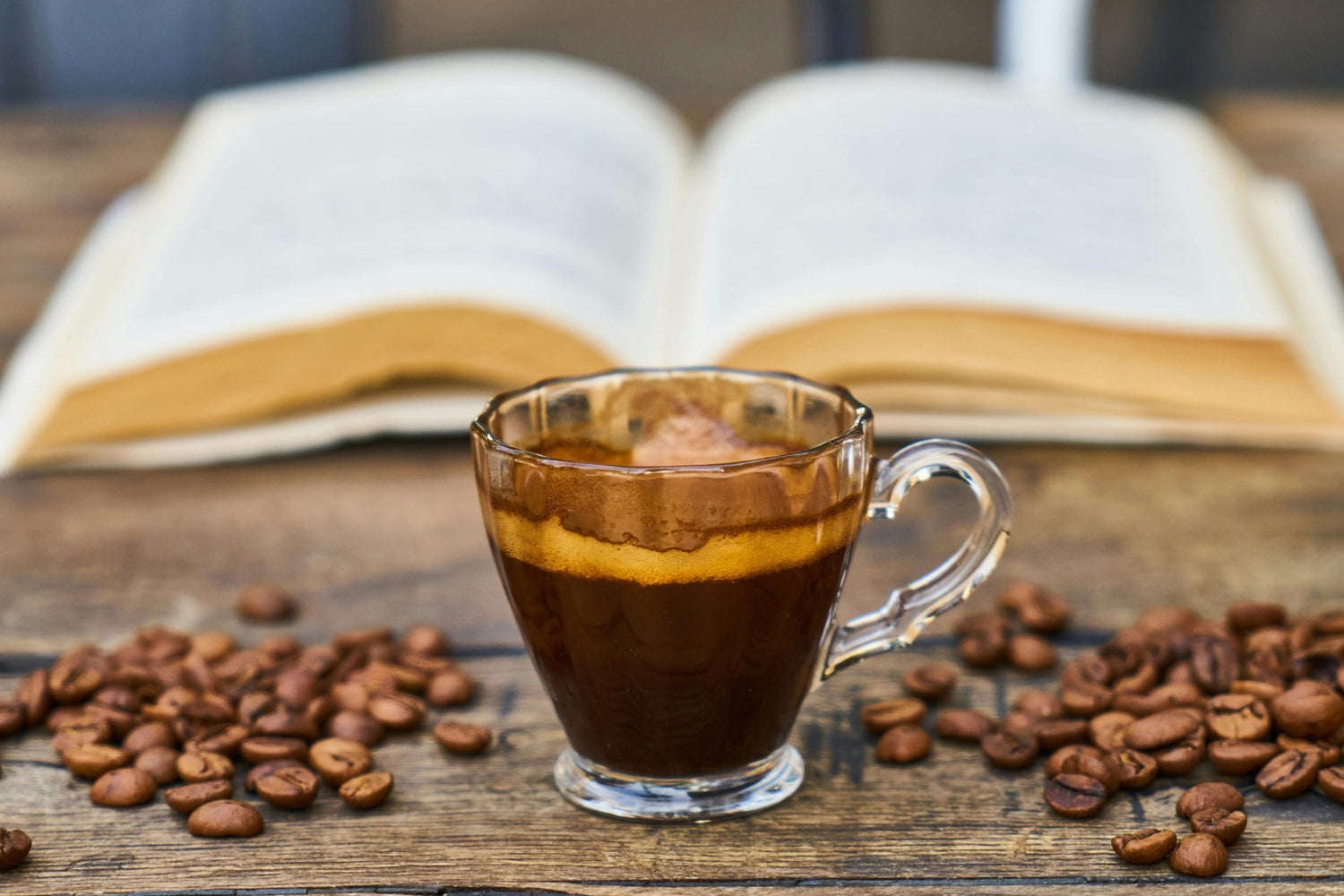 Clear glass mug filled with coffee on a wooden surface with scattered coffee beans and an open book in the background.