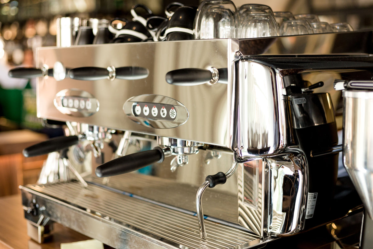 A close-up of a professional espresso machine with a polished stainless steel body, black handles, and glass carafes visible in the background.