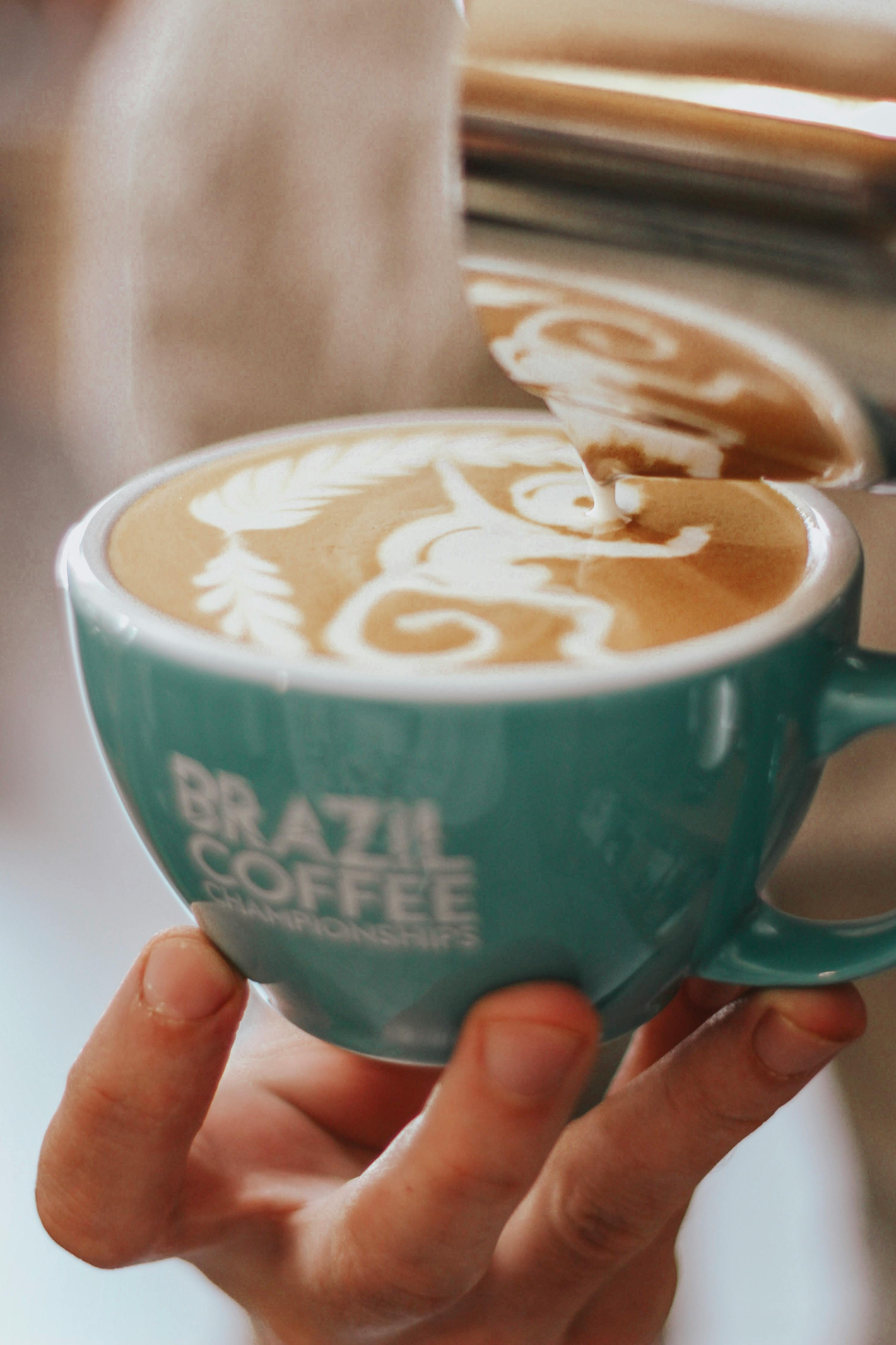 Teal mug with 'Brazil Coffee' branding holding a cappuccino with latte art.