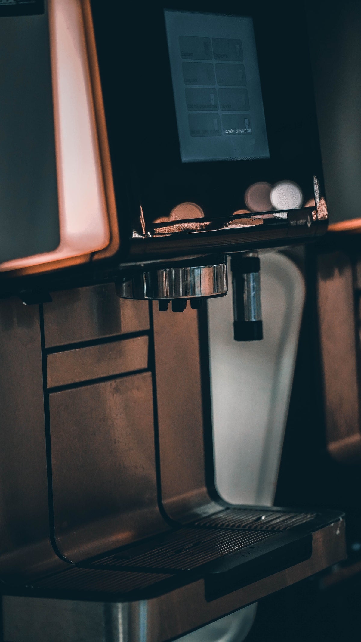 A close-up, angled view of a modern coffee machine with a metallic finish, featuring a digital display screen on top, a drip tray, and a steam wand, set against a dark background with warm ambient lighting.