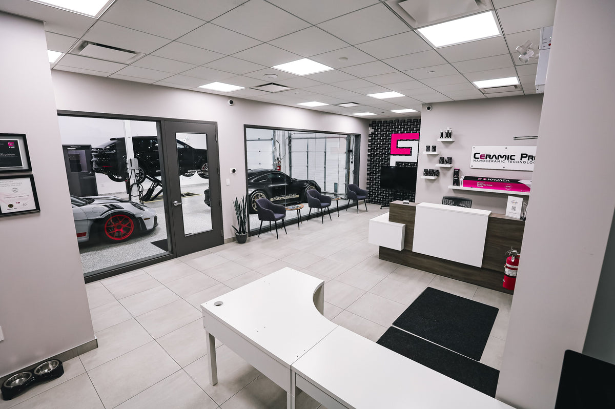 A modern office reception area with a white desk, a white and wood counter, black chairs, large windows, and a visible sports car outside through the glass.