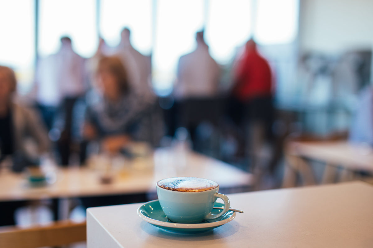 A light blue ceramic coffee cup and saucer filled with cappuccino sits on a table in a cafe, with blurred people sitting and standing in the background.