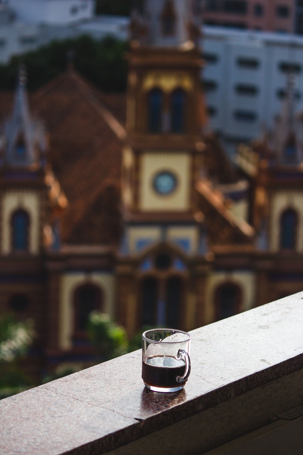 A small clear glass cup filled with dark coffee sits on a stone balcony ledge, with a blurred historic church building in the background.