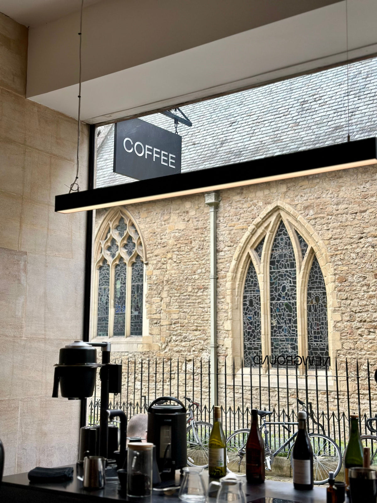 A modern black linear pendant light with a warm LED glow hangs above a coffee station. Through a large window, a historic stone building with Gothic arched windows is visible, along with a bicycle and the word 'COFFEE' on the sign.