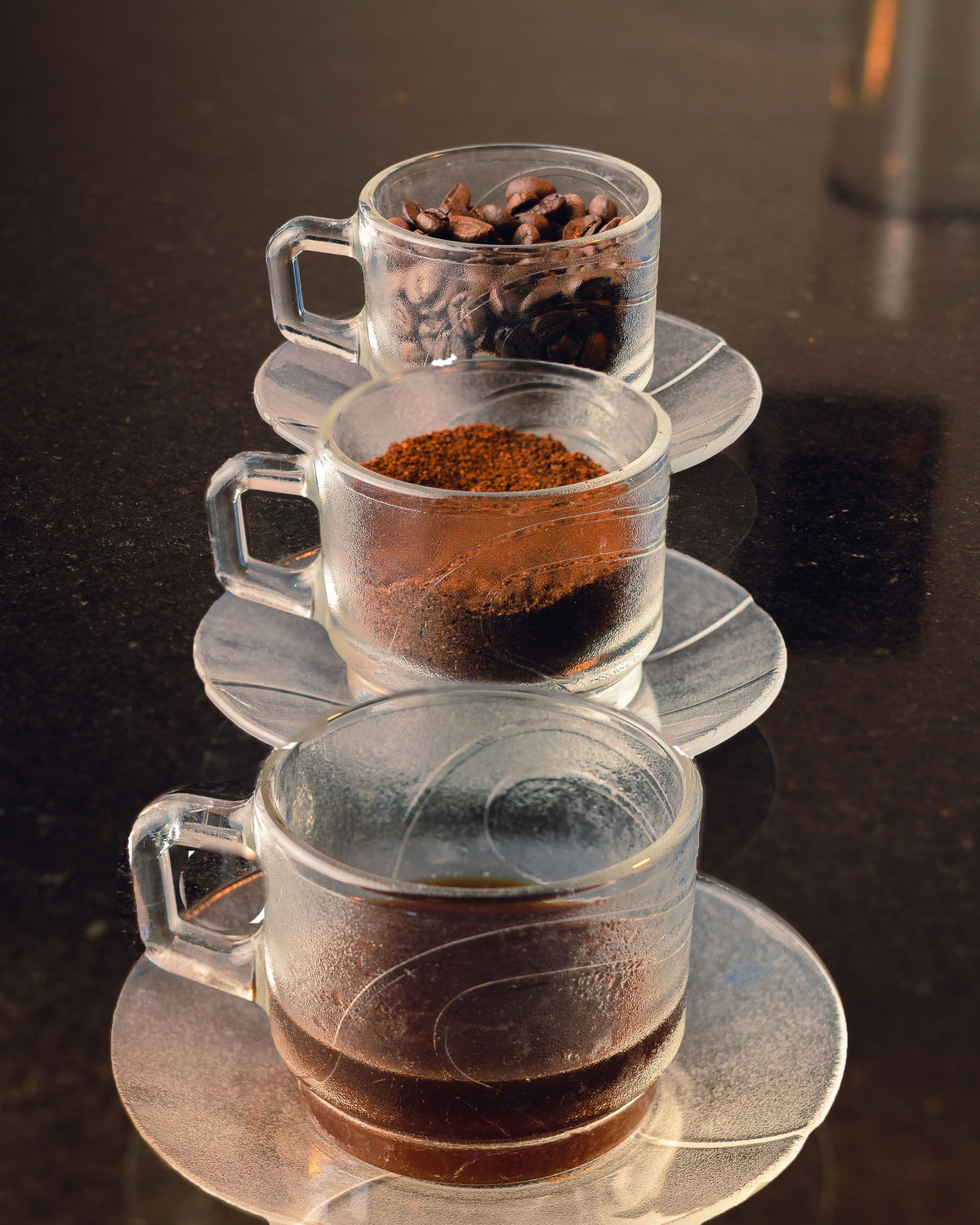 Three clear glass coffee cups with saucers stacked vertically on a dark countertop, containing whole coffee beans, ground coffee, and brewed coffee respectively.
