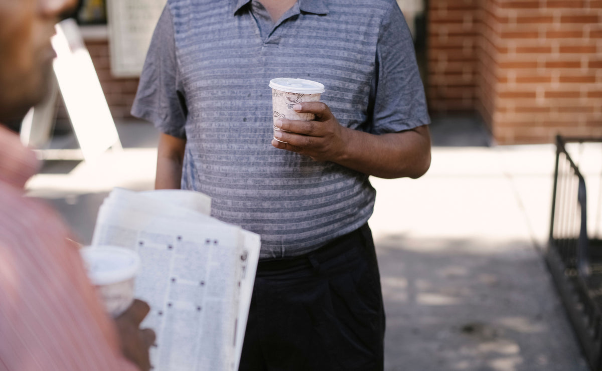 A man in a gray striped short-sleeve polo shirt and black pants stands on a sidewalk holding a paper coffee cup, with another person partially visible in the foreground holding a Bible