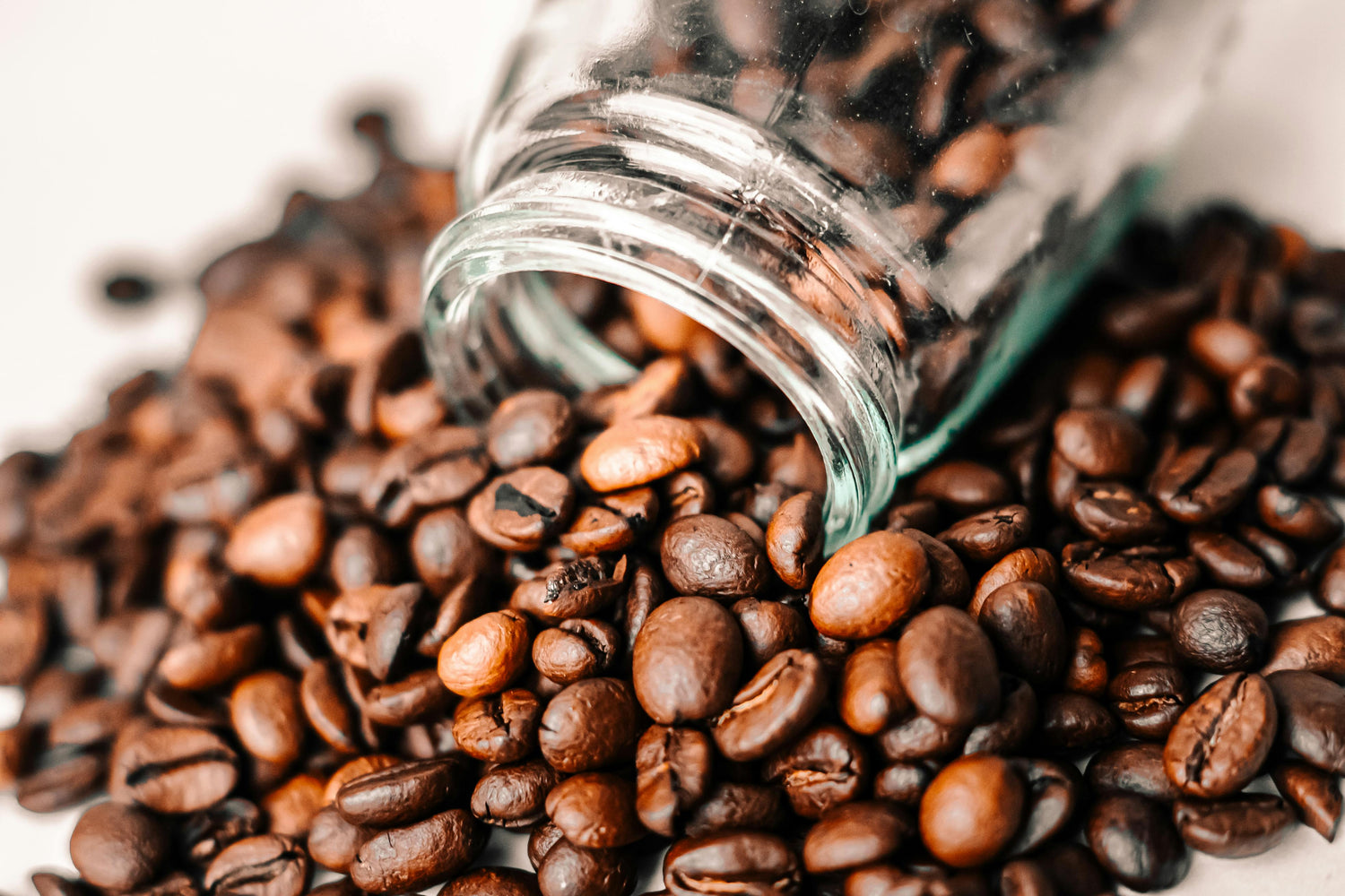 Glass jar tipped over with coffee beans spilling out on a light background