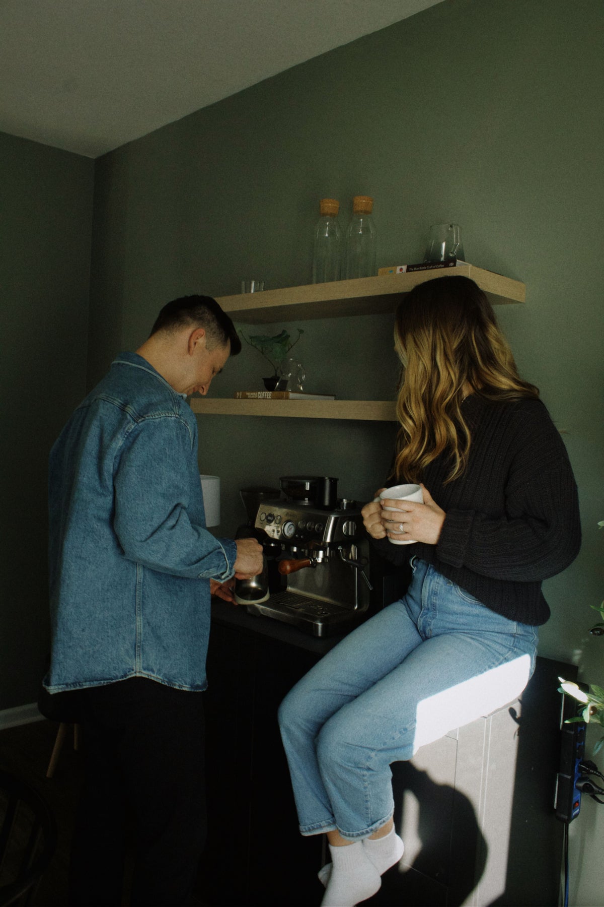 A man in a blue denim shirt stands beside a coffee machine while a woman in a black sweater and light blue jeans sits on a white stool holding a white mug in a dimly lit room with dark green walls and floating shelves.