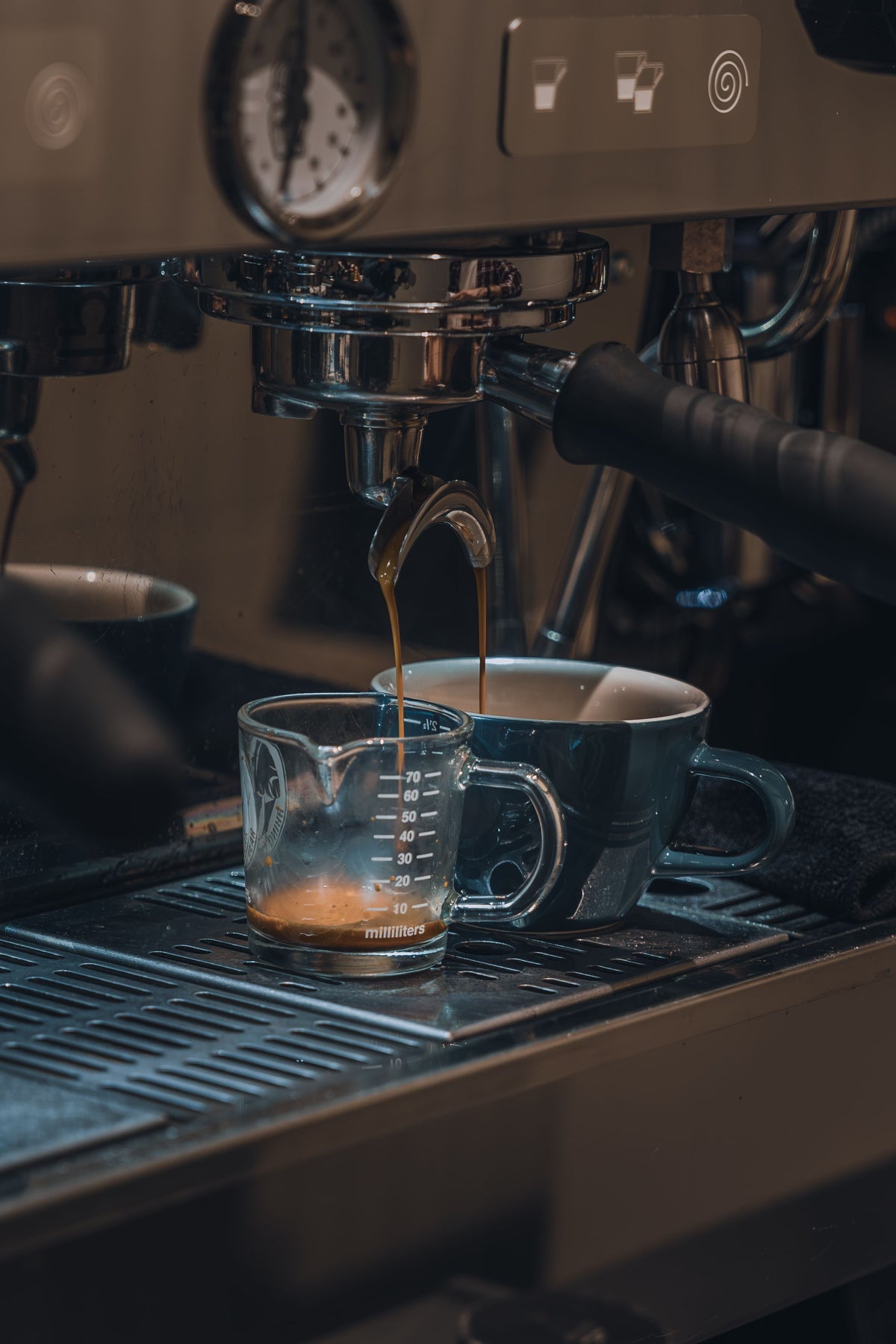 Espresso being poured from a commercial espresso machine into a clear glass measuring cup and a dark ceramic mug on a drip tray.